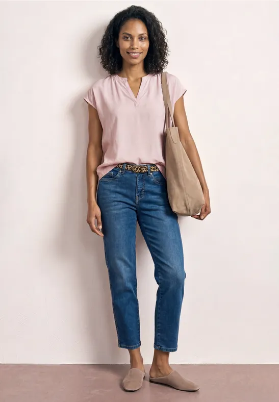 Vrouw in roze blouse, blauwe spijkerbroek, bruine loafers en stoffen tas.