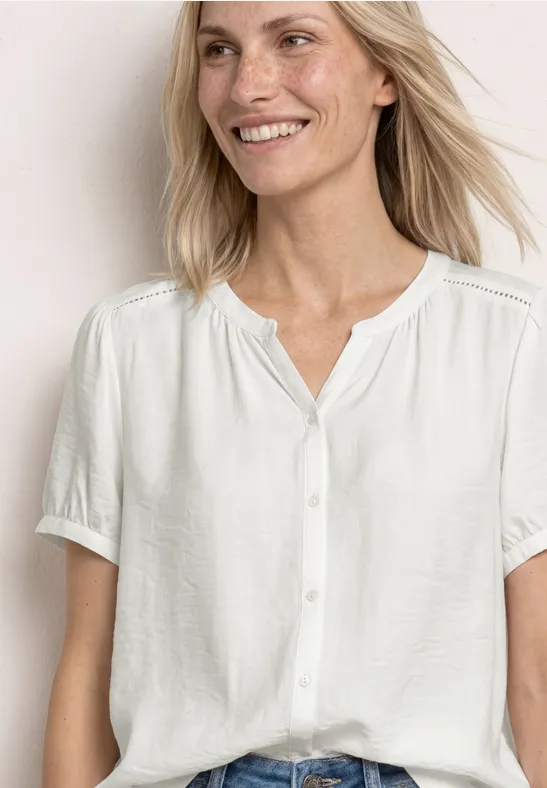 Een vrouw met lichtblond haar draagt een witte blouse met korte mouwen en knopen.