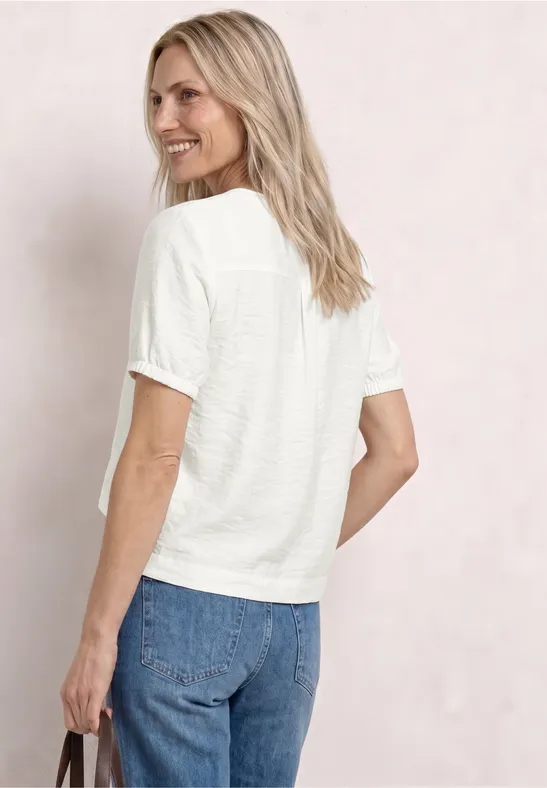Vrouw van achter getoond in wit T-shirt en blauwe jeans, handtas in hand, los blond haar, neutrale achtergrond.