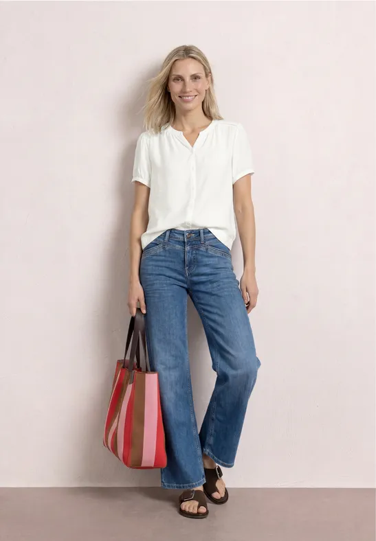 Vrouw in wit overhemd, blauwe jeans en gestreepte tas poseert voor lichte achtergrond.