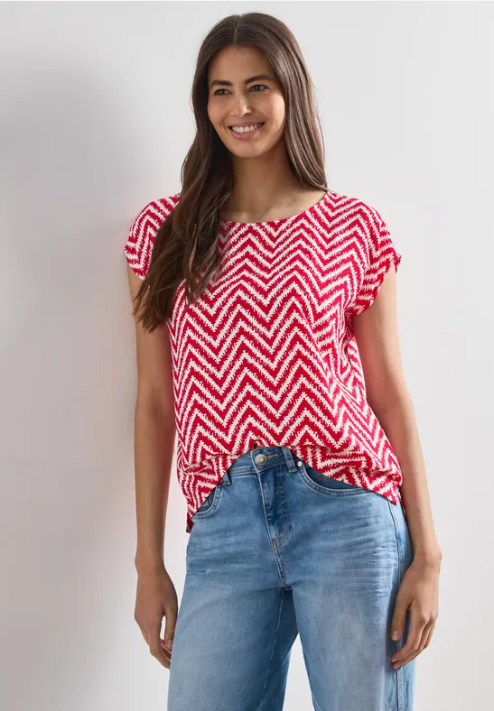 Een vrouw draagt een rood-wit zigzagshirt en een blauwe spijkerbroek tegen een lichte achtergrond.