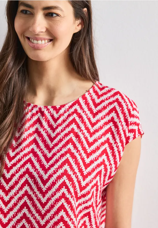Glimlachende vrouw in een rood-wit zigzagpatroon top met korte mouwen.