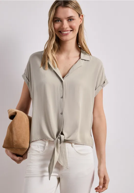 Frau in kurzärmligem Hemd vorne geknotet, weißer Hose und beiger Clutch.