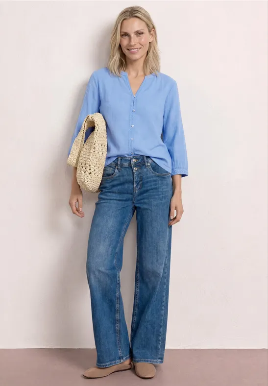 Frau in hellblauer Bluse, weitem Jeans und Tragetasche aus geflochtenem Stroh vor hellem Hintergrund.