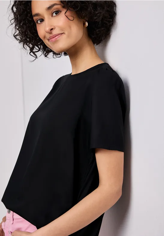 Person steht seitlich an einer Wand, trägt eine schwarze Bluse mit kurzen Ärmeln und rosa Hose.