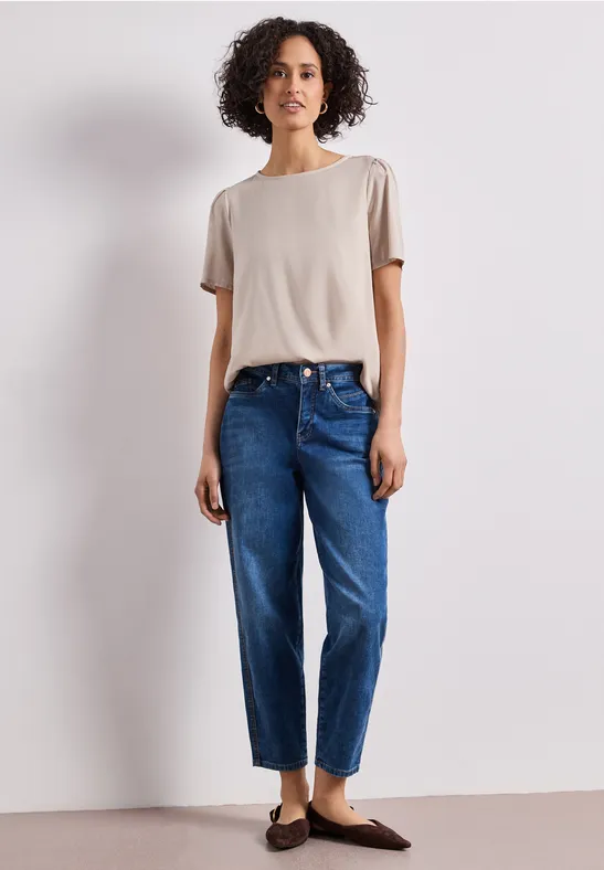 Persoon in beige T-shirt, blauwe jeans en bruine loafers, staand voor een witte achtergrond.