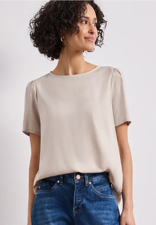 Een vrouw draagt een crème korte mouwen blouse en blauwe jeans tegen een lichte achtergrond.