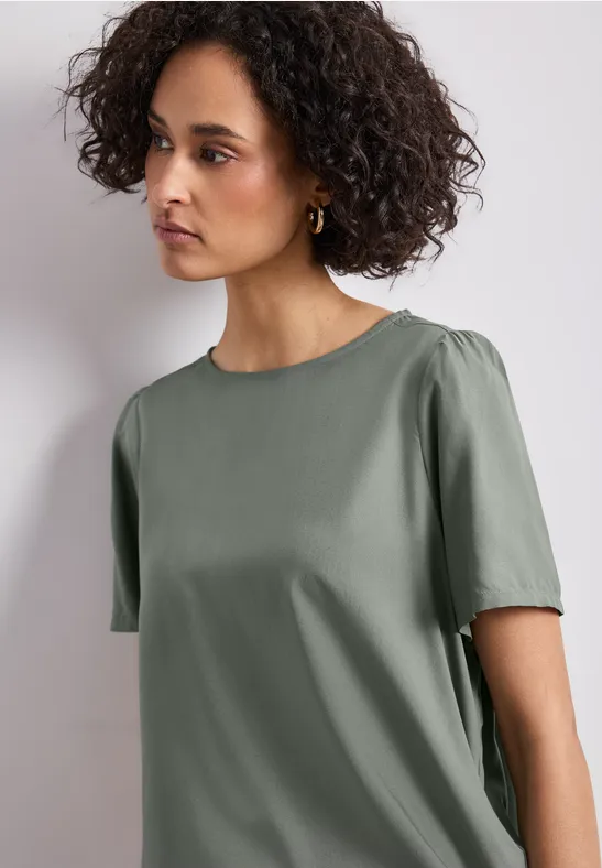 Een vrouw met krullend haar draagt een eenvoudig kort olijfgroen T-shirt voor een lichte achtergrond.