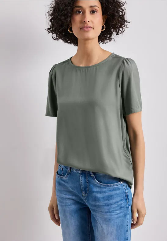 Persoon draagt groen shirt met korte mouwen en blauwe spijkerbroek voor lichte achtergrond.