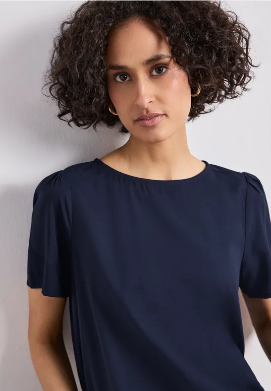 Eine Person mit lockigem Haar trägt eine dunkelblaue, kurzärmelige Bluse vor weißem Hintergrund.