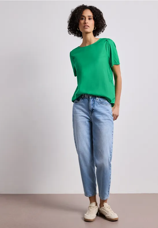 Une personne porte un t-shirt vert, un jean clair et des baskets, debout sur fond neutre.