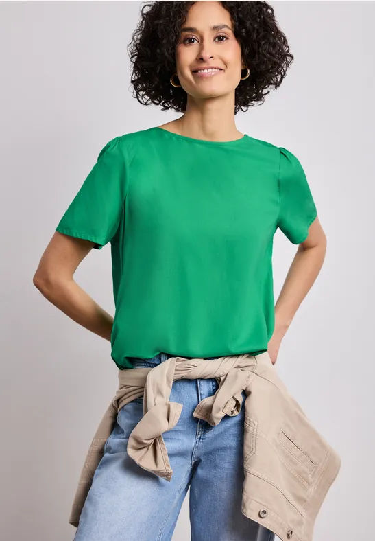Personne aux cheveux bouclés porte un t-shirt vert, un jean et une veste beige nouée à la taille sur fond neutre.