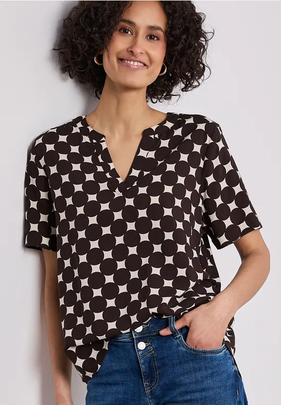 Femme porte une blouse noir et blanc à manches courtes, motif cercles et carrés, partiellement rentrée dans un jean.