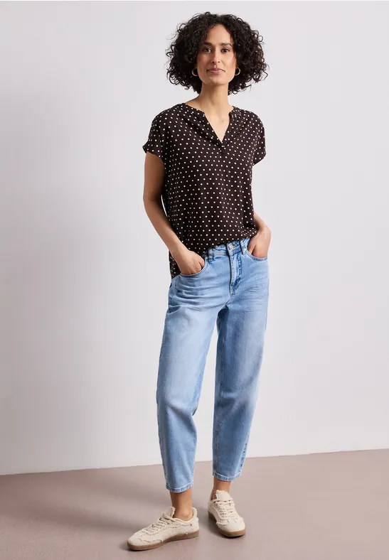 Eine Frau mit kurz geschnittener lockiger Frisur trägt ein schwarzes Polka-Dot-Shirt, blaue Jeans und weiße Sneakers.