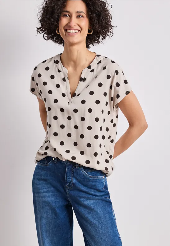 Vrouw in blauwe jeans en beige blouse met zwarte stippen en korte mouwen, handen achter de rug.