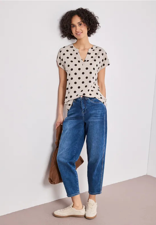 Vrouw in een stippenblouse, blauwe jeans en witte sneakers leunt nonchalant tegen een muur.