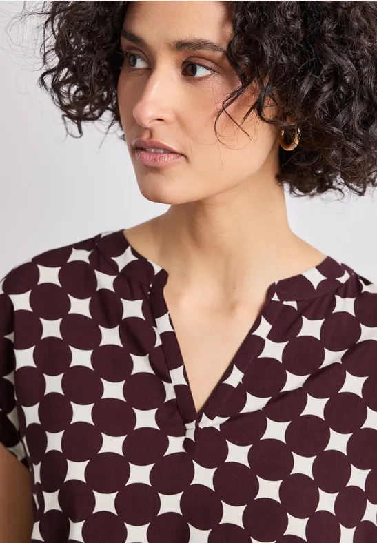 Porträt einer Frau mit dunklen Locken in einem weißen Top mit großen dunkelbraunen Punkten.