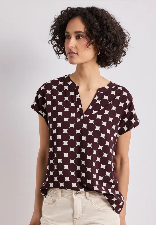 Frau mit lockigem Haar trägt eine kurzärmelige Bluse mit geometrischem Muster und eine helle Hose.