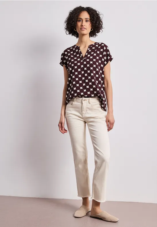 Eine Frau mit dunklem, lockigem Haar trägt eine schwarze Bluse mit weißem Muster, helle Hose und Pantoffeln.