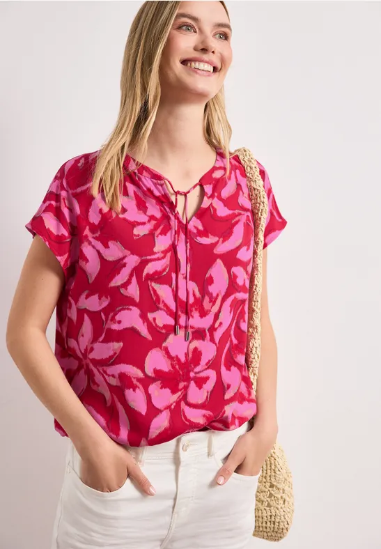 Een vrouw draagt een roze top met bloemmotief, een lichte broek en houdt een strotas vast.