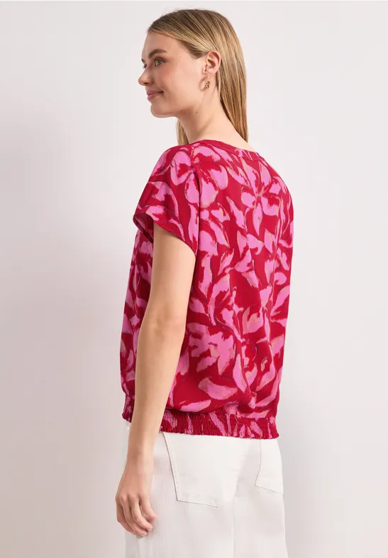 Vrouw van opzij in een roze top met bladmotief en witte broek.