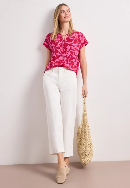 Model draagt roze shirt met print, witte culotte-jeans, beige loafers en een gevlochten strooien tas.