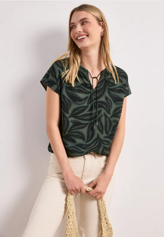 Vrouw draagt donkergroene blouse met bladmotief, witte broek en houdt een gevlochten tas vast.