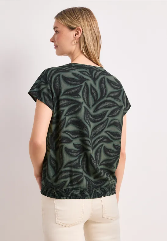 Achteraanzicht van een vrouw in groene bladprint blouse met korte mouwen en lichte broek voor witte achtergrond.