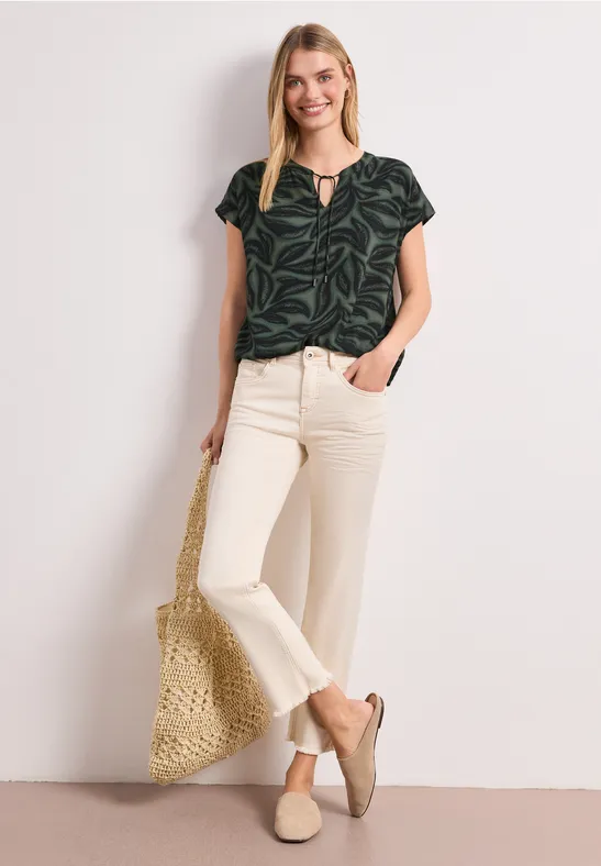Vrouw draagt een donkergroen blouse met bladprint, beige uitlopende broek, platte schoenen en een strooien tas.