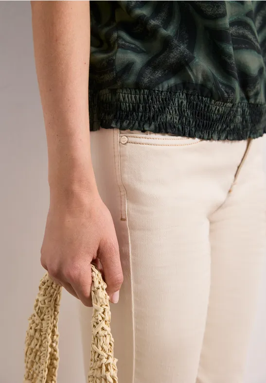 Persoon in beige broek en groen patroon-top houdt een gevlochten tas in de hand.