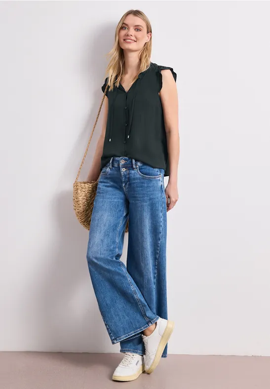 Vrouw in zwarte mouwloze top, wijde spijkerbroek, witte sneakers met rieten tas voor een lichte muur.