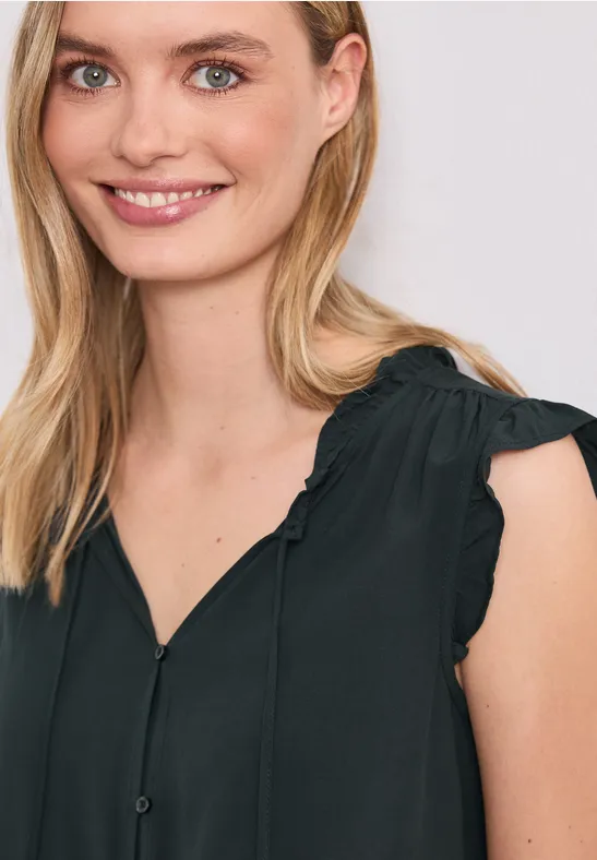 Een vrouw draagt een mouwloze zwarte blouse met ruches op de schouders.