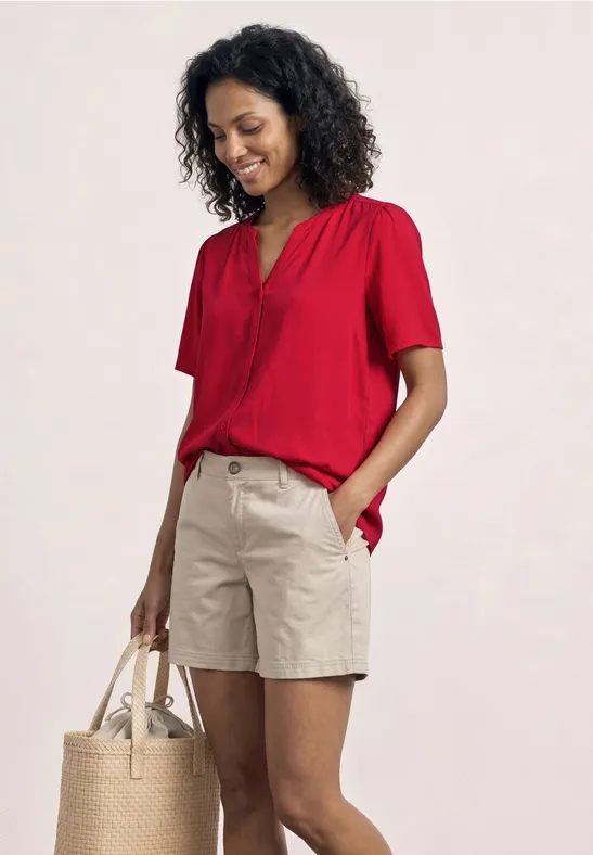 Een persoon draagt een rood shirt met korte mouwen, beige shorts en houdt een strooien tas vast.
