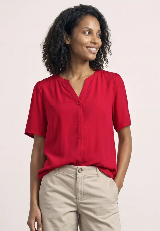 Een vrouw met krullend haar draagt een rood bloesje en een lichte broek.