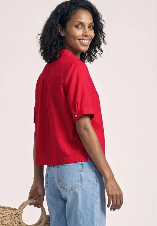 Vrouw in zijaanzicht met krullend haar, in een rood kort shirt en blauwe jeans, met een tas in de hand.
