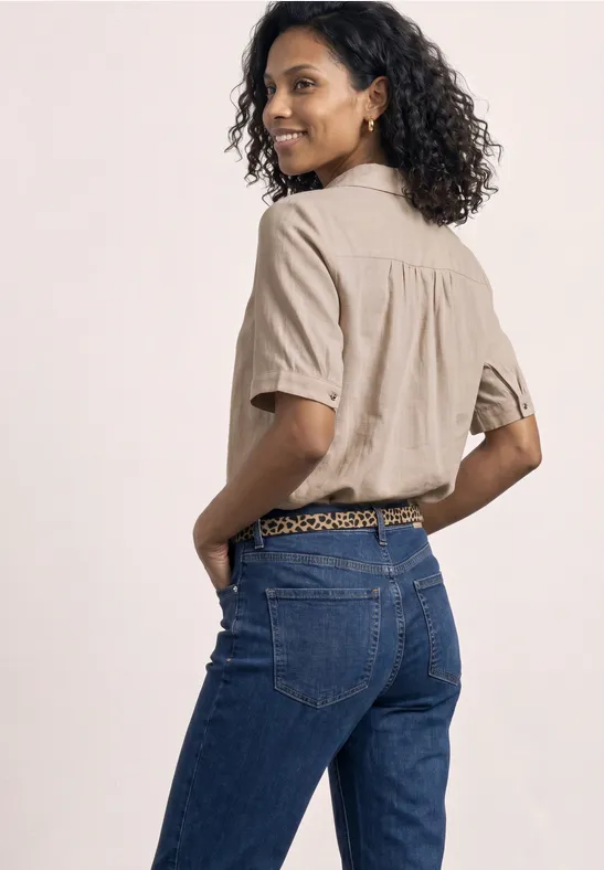Vrouw met krullend haar in een beige overhemd en blauwe jeans, handen in de zakken, ruggezicht te zien.