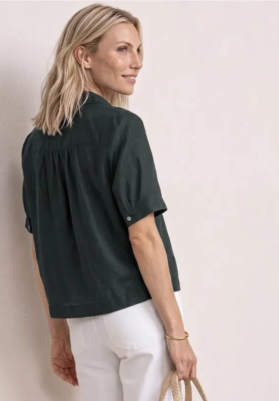 Personne vue de côté portant une chemise noire et un pantalon blanc, tenant une pochette, regard détourné.