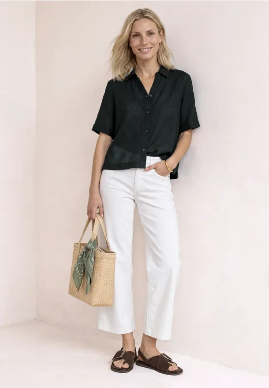 Personne blonde en chemise noire, pantalon blanc, sandales brunes et sac en paille devant un mur blanc