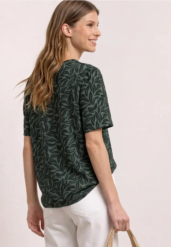 Vrouw van achteren in groen t-shirt met bladprint en witte broek tegen neutrale achtergrond.