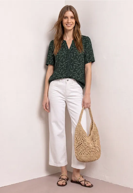 Vrouw in groene top, witte broek, zwarte sandalen en beige gehaakte tas.