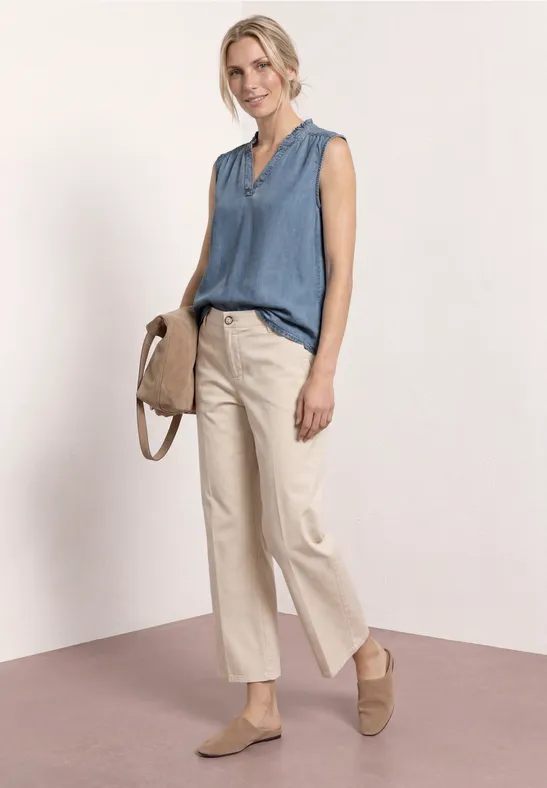 Een vrouw in casual outfit: mouwloze blauwe blouse, beige wijde broek, beige instappers en bijpassende tas.