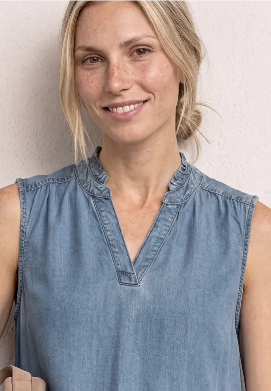 Vrouw in mouwloze denimtop met ruches bij de hals voor een lichte muur.