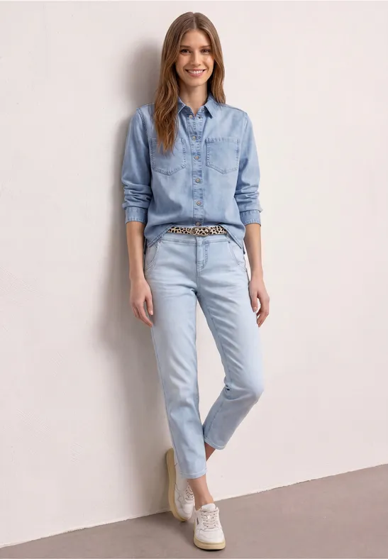 Vrouw leunt tegen een lichtroze muur, draagt lichtblauwe jeans, een denimblouse, een dessinriem en witte sneakers.