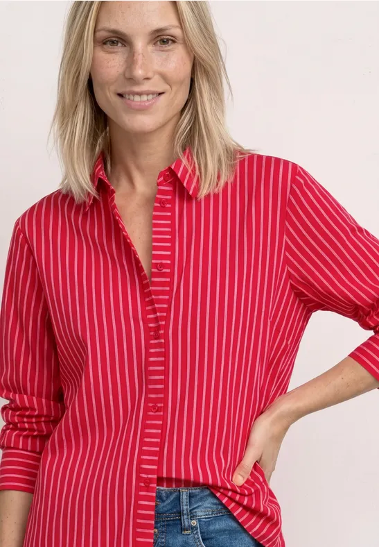 Blonde Frau trägt rotes Hemd mit weißen Streifen, lächelt selbstbewusst und hat eine Hand in der Hüfte.