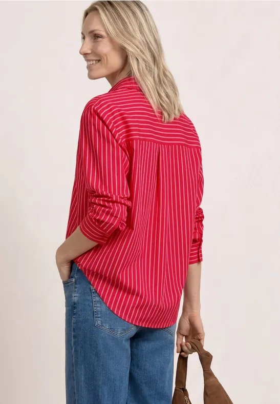 Rückansicht: Frau in rot-weiß gestreiftem Hemd, blauer Jeans, Händen in Taschen, braune Tasche in der Hand.