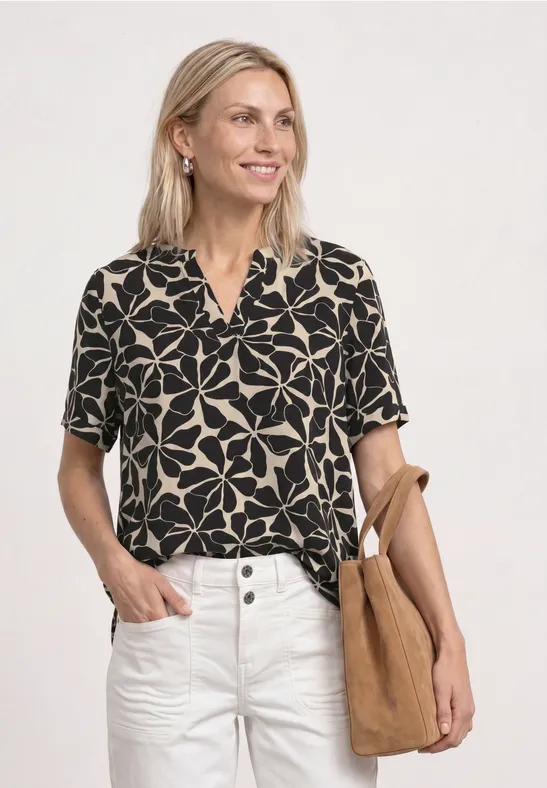 Vrouw in zwarte bloes met wit bloemenpatroon, witte broek en beige tas tegen witte achtergrond.