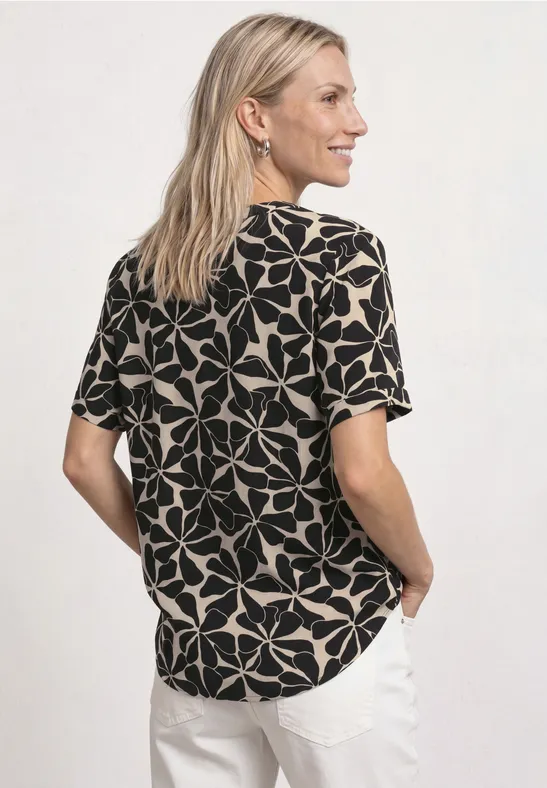 Vrouw van opzij met blik over schouder, draagt zwart-beige bloes met patroon en witte broek.