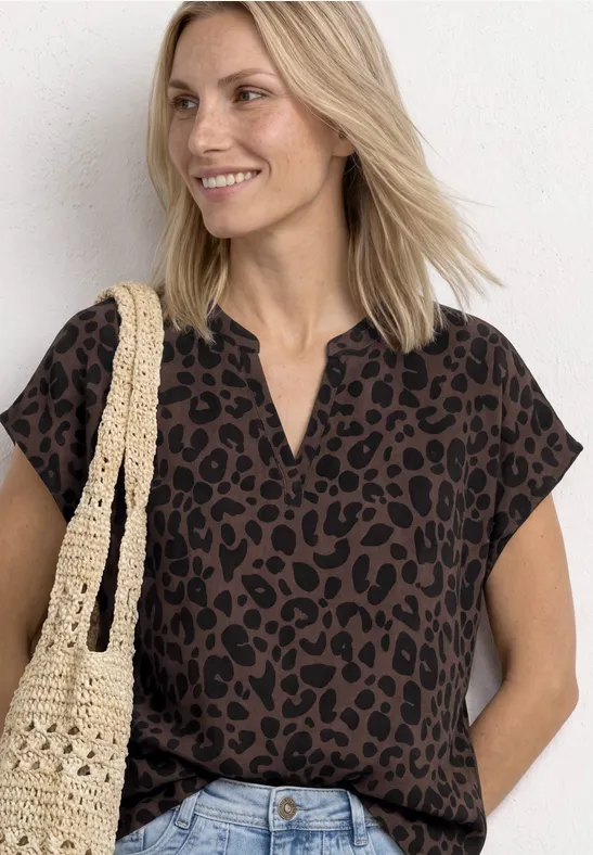 Femme en haut imprimé léopard, jean et sac au crochet, posture décontractée devant un mur blanc.