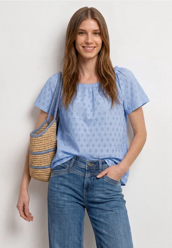 Vrouw draagt lichtblauw kort mouwshirt met patroon, spijkerbroek en rieten schoudertas.