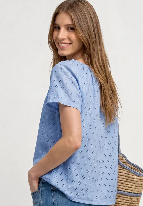 Vrouw in zijaanzicht met lichtblauw, geborduurd kort mouw topje en een strooien tas in haar hand.